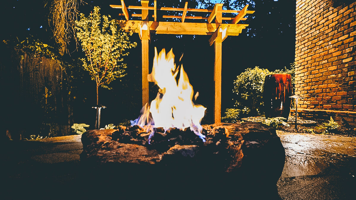fire burning in stone fire pit in front of wooden backyard arbor
