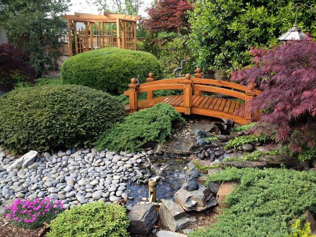 wooden bridge crossing over stone garden fountain with shrubberies and softscaping next to pergola